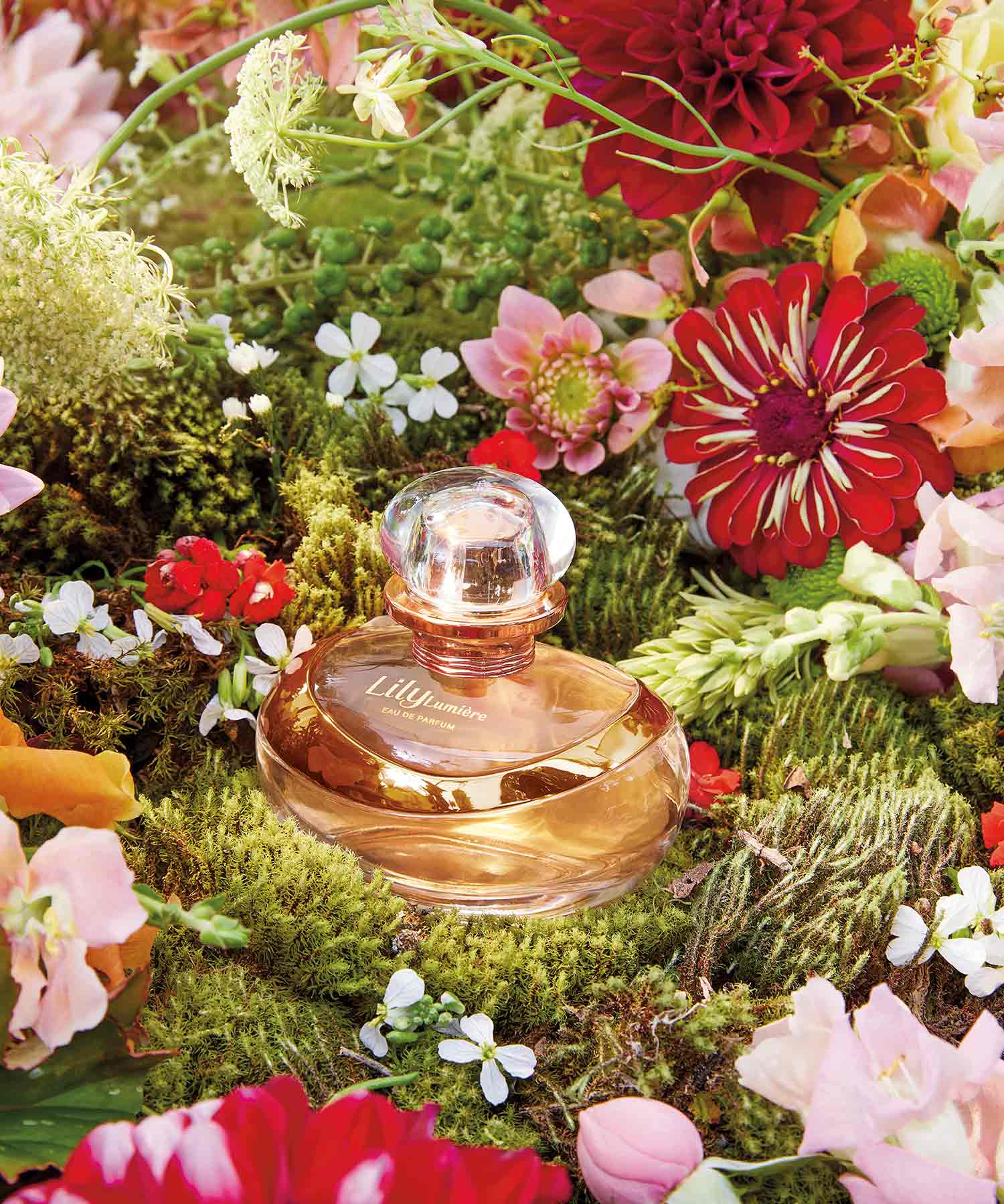 Frasco de perfume Lily num ambiente floral alusivo ao Dia da Mãe. A embalagem é em vidro transparente, com um líquido amarelo claro dentro e uma tampa dourada. O rótulo tem o nome do perfume em letras elegantes.